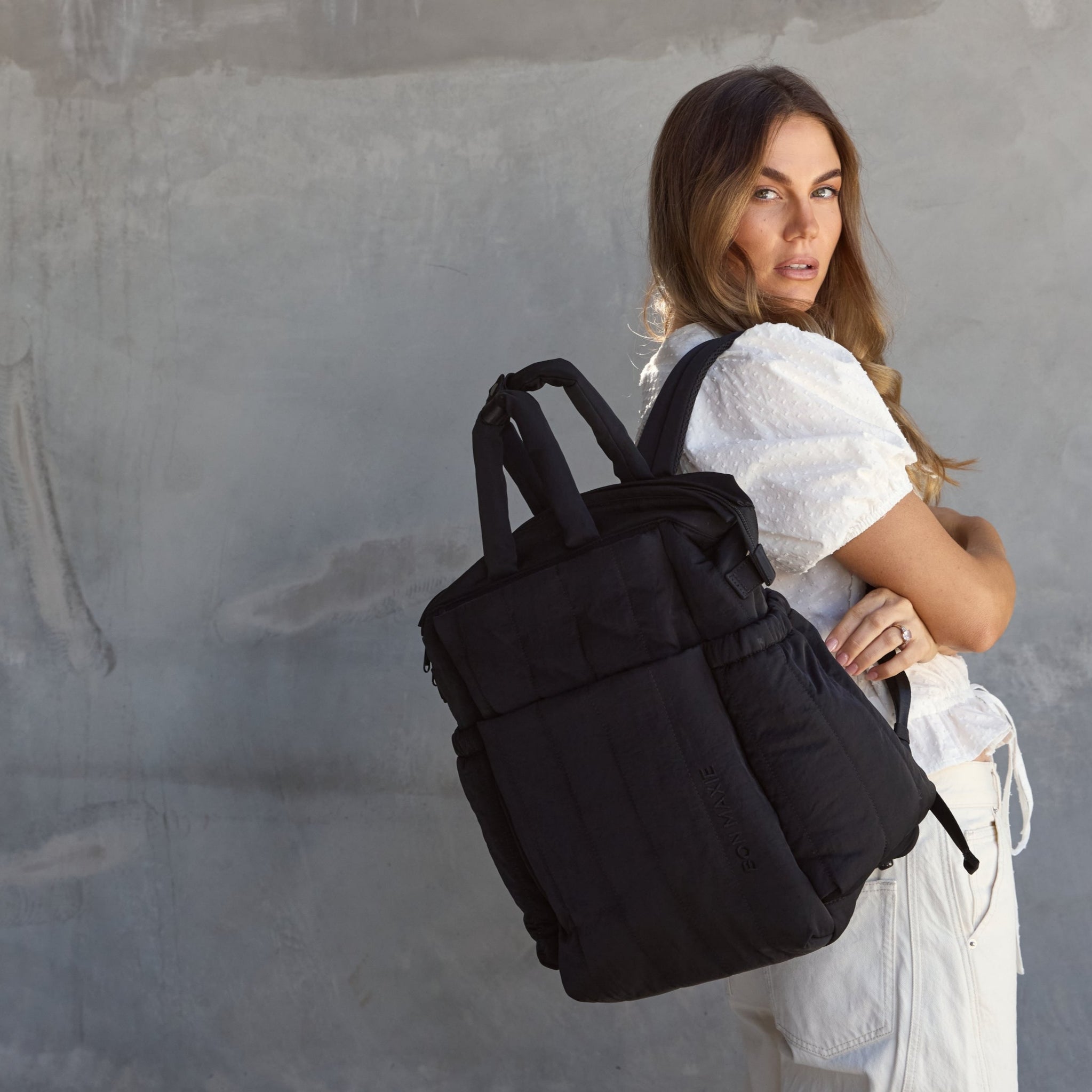 Woman with a black backpack standing against a gray wall.