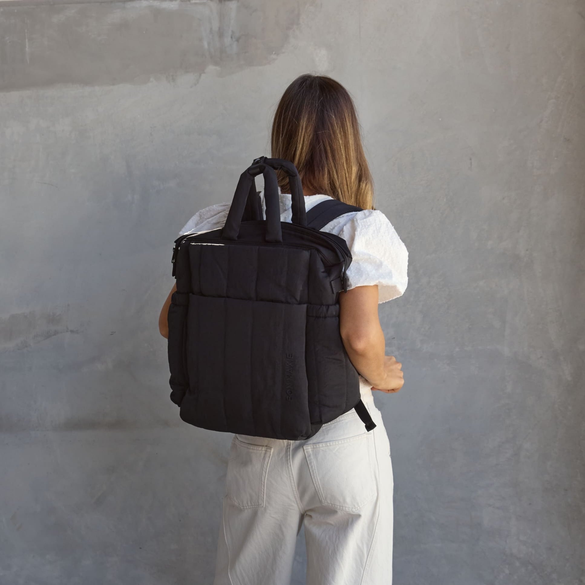Person wearing a black backpack in an indoor setting with a concrete wall and window.