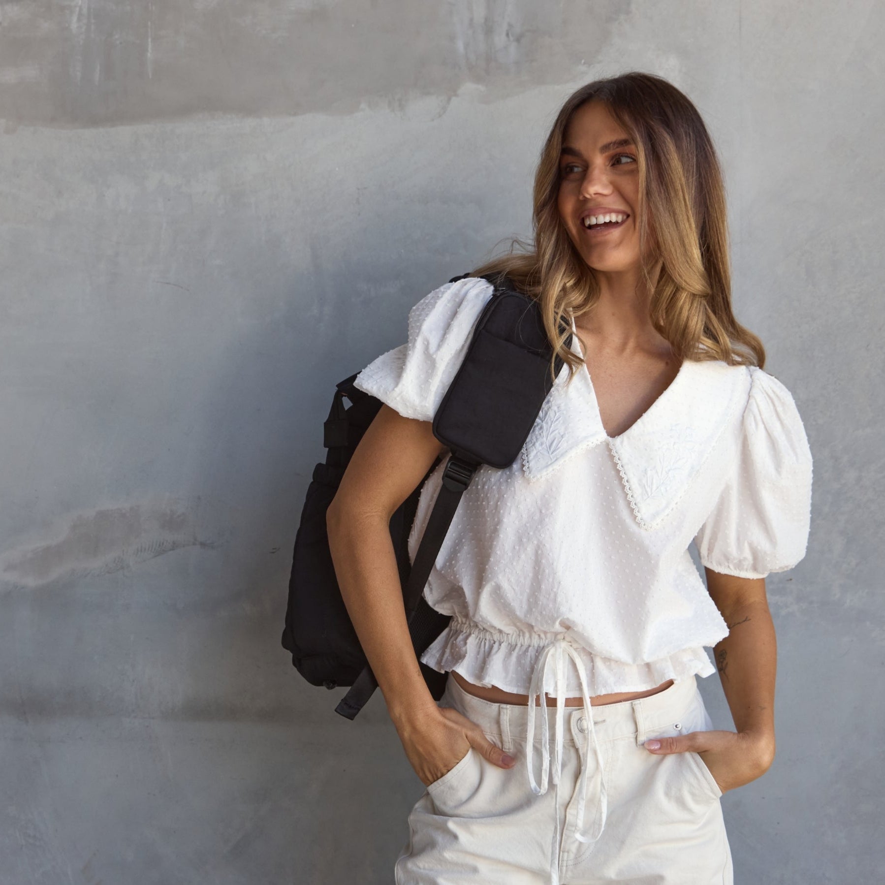 Woman in a white outfit with a black backpack standing against a gray wall.