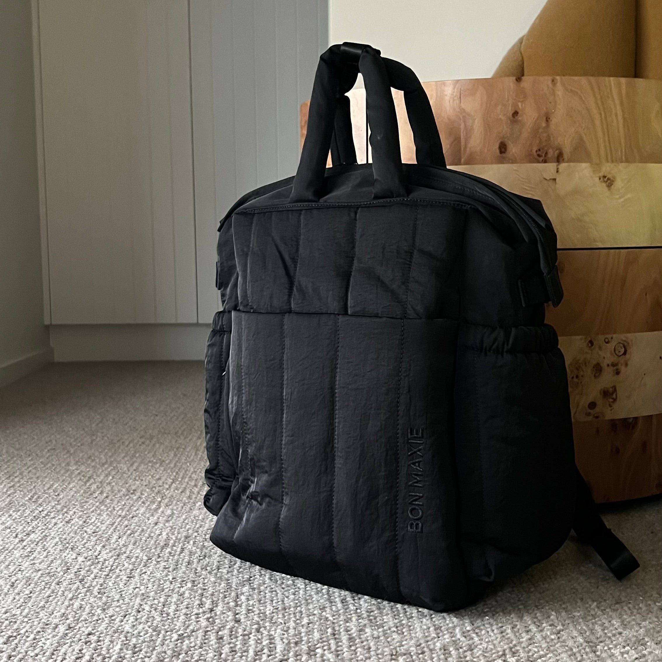 Black backpack on a carpeted floor with a neutral-colored wall and furniture in the background