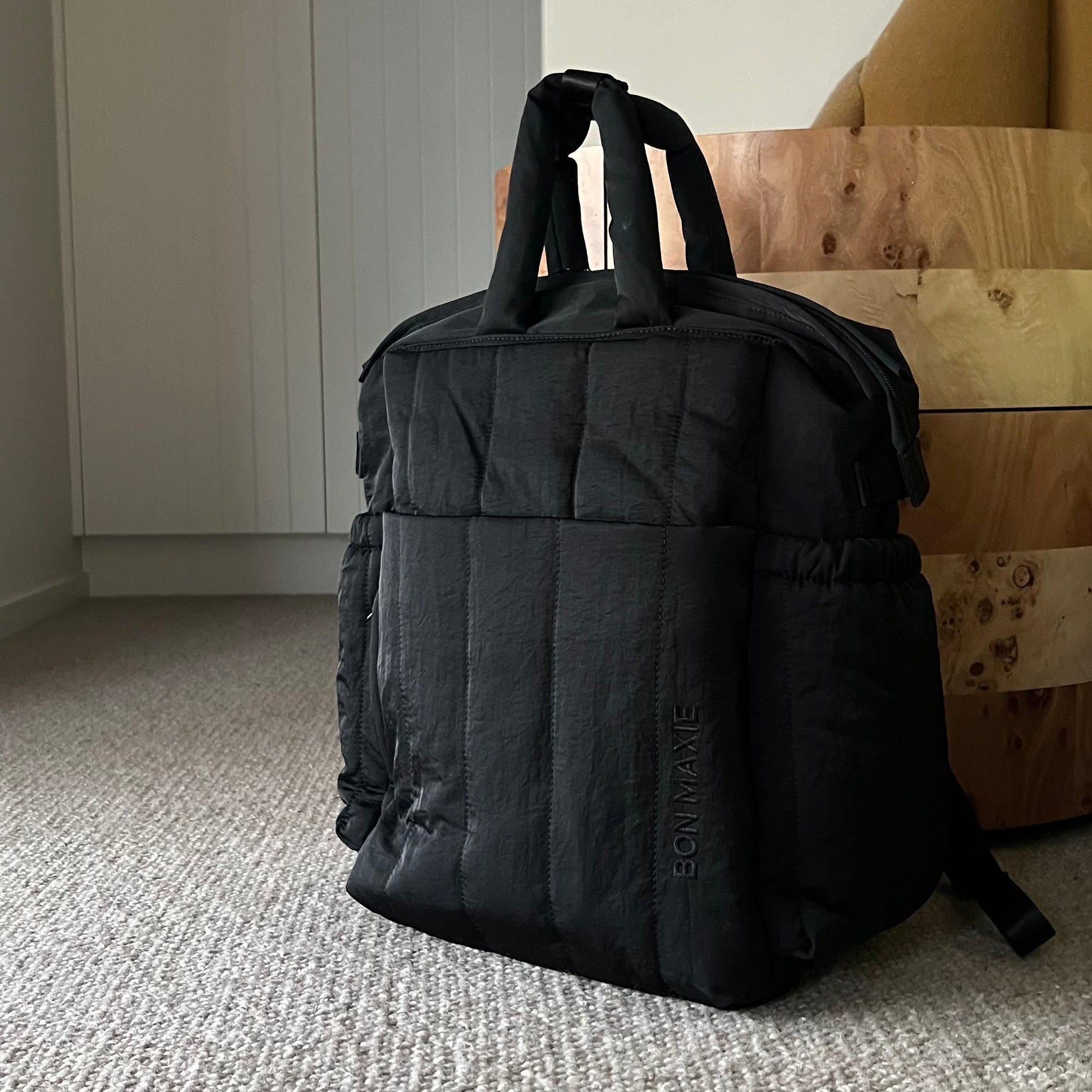 Black backpack on a carpeted floor with a neutral-colored wall and furniture in the background