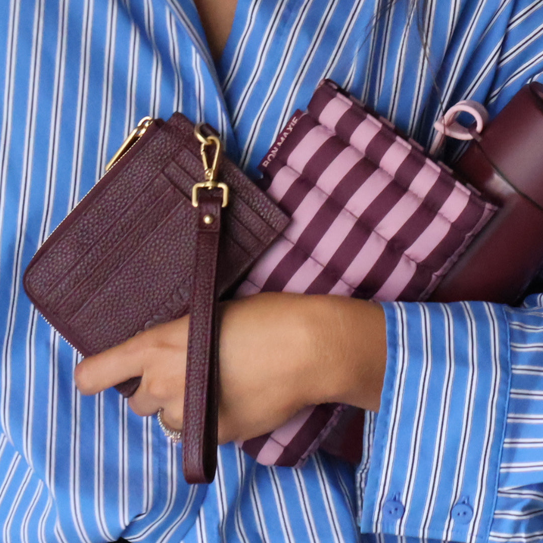 A person in a blue and white striped shirt holding three small leather accessories: a dark burgundy textured wristlet with gold hardware, a pink and burgundy striped zip pouch, and a dark burgundy shoulder bag.